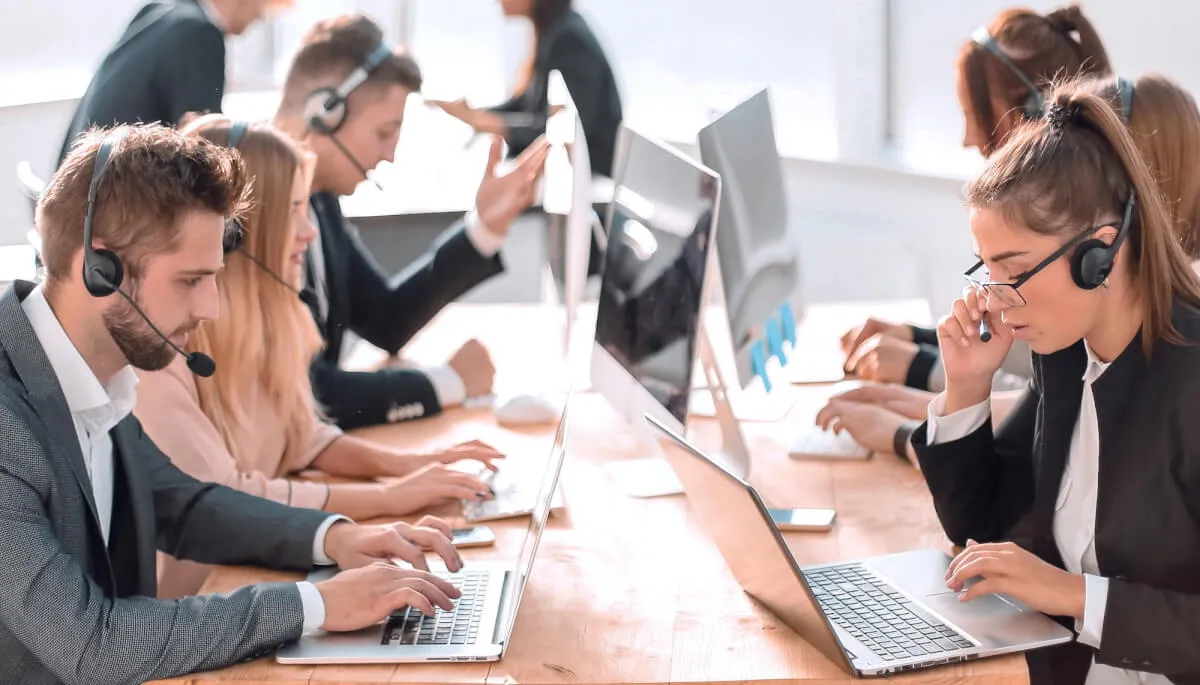 Agentes de un contact center con auriculares trabajando con portátiles en una larga mesa compartida
