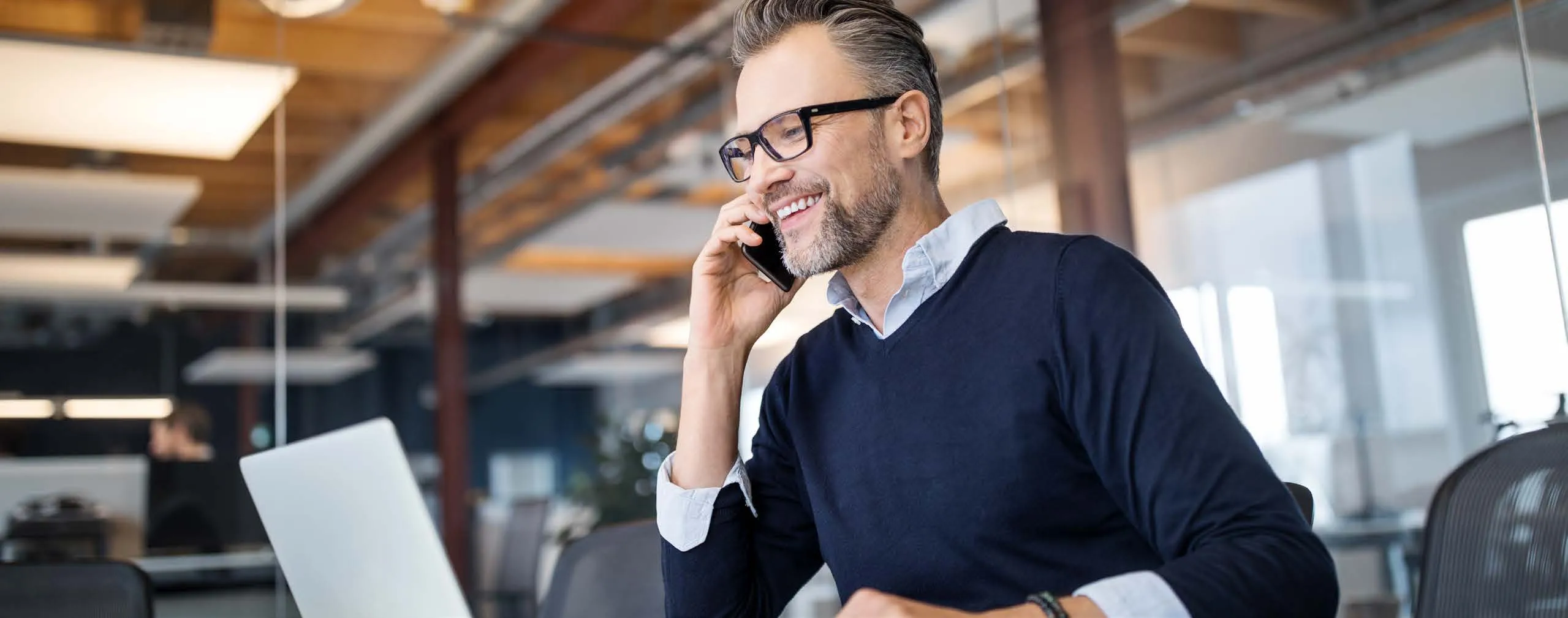 Un hombre con gafas sonríe mientras habla por teléfono en una mesa de una oficina luminosa, con un portátil delante.