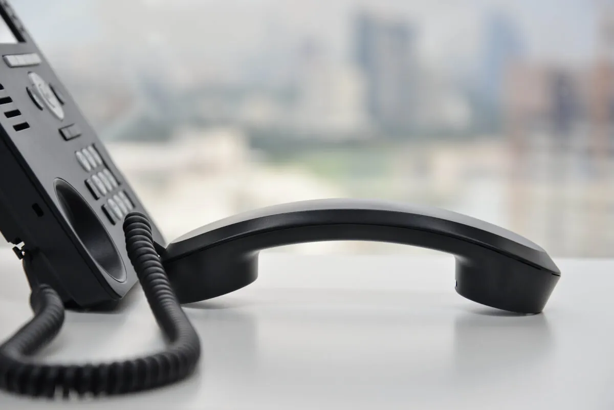 Teléfono fijo de oficina negro con el auricular apoyado sobre un escritorio blanco y una ciudad desenfocada al fondo